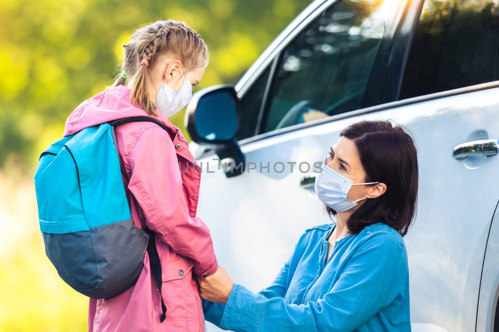 Schoolgirl with mother in protective masks at car parking before classes during coronavirus pandemic