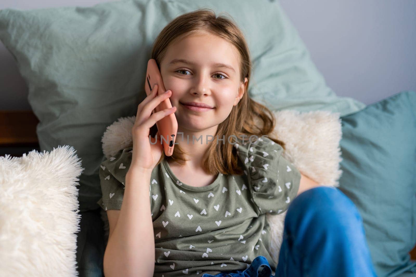 Pretty preteen girl sitting in the bed and talking by smartphone at home in the bedroom. Beautiful kid with cell phone communicating indoors