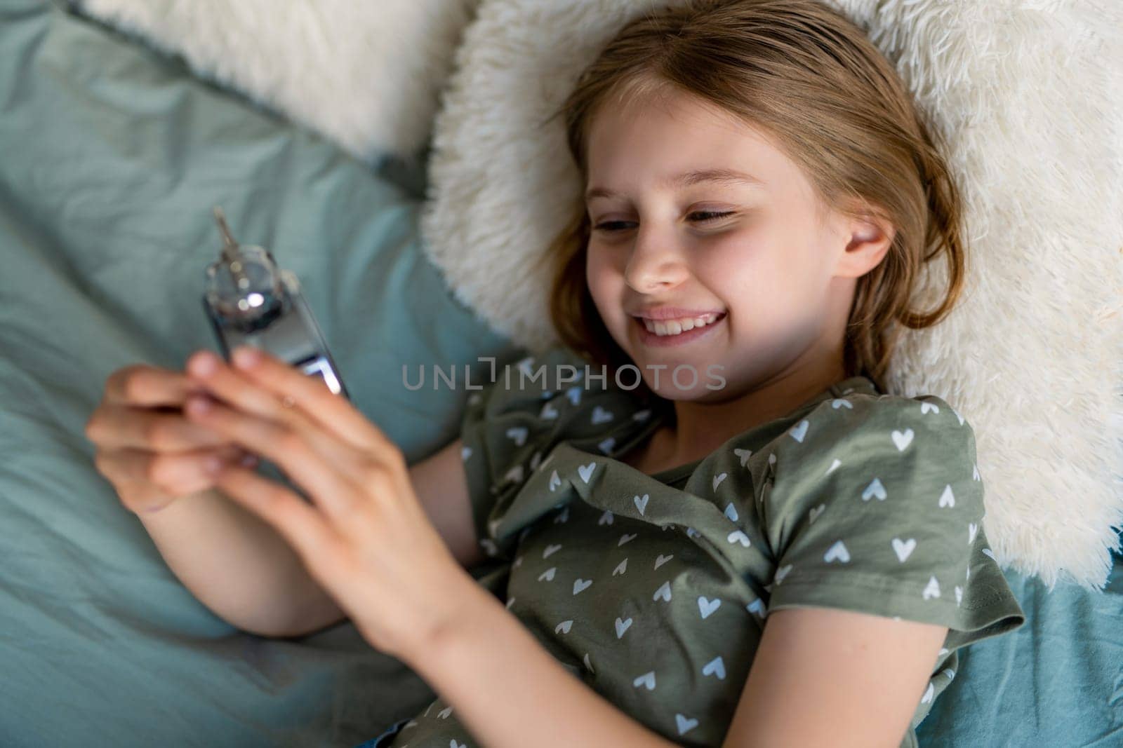 Portrait of preteen beautiful blond hair girl lying in the bed with clock. Female kid schoolgirl smiling in the bedroom