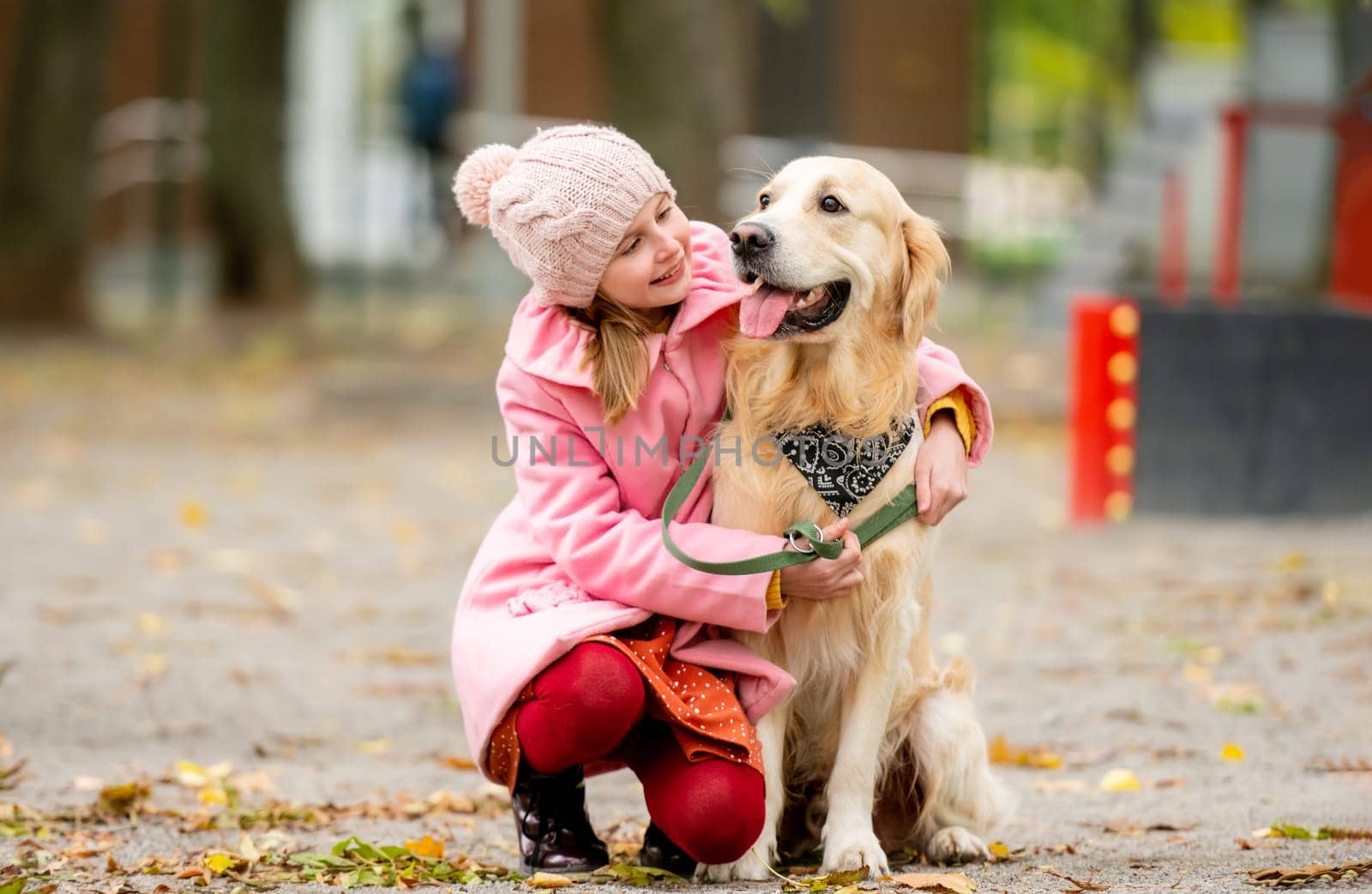Preteen girl kid with golden retriever dog sitting in the park, smiling and petting doggy. Female child with pet at autumn