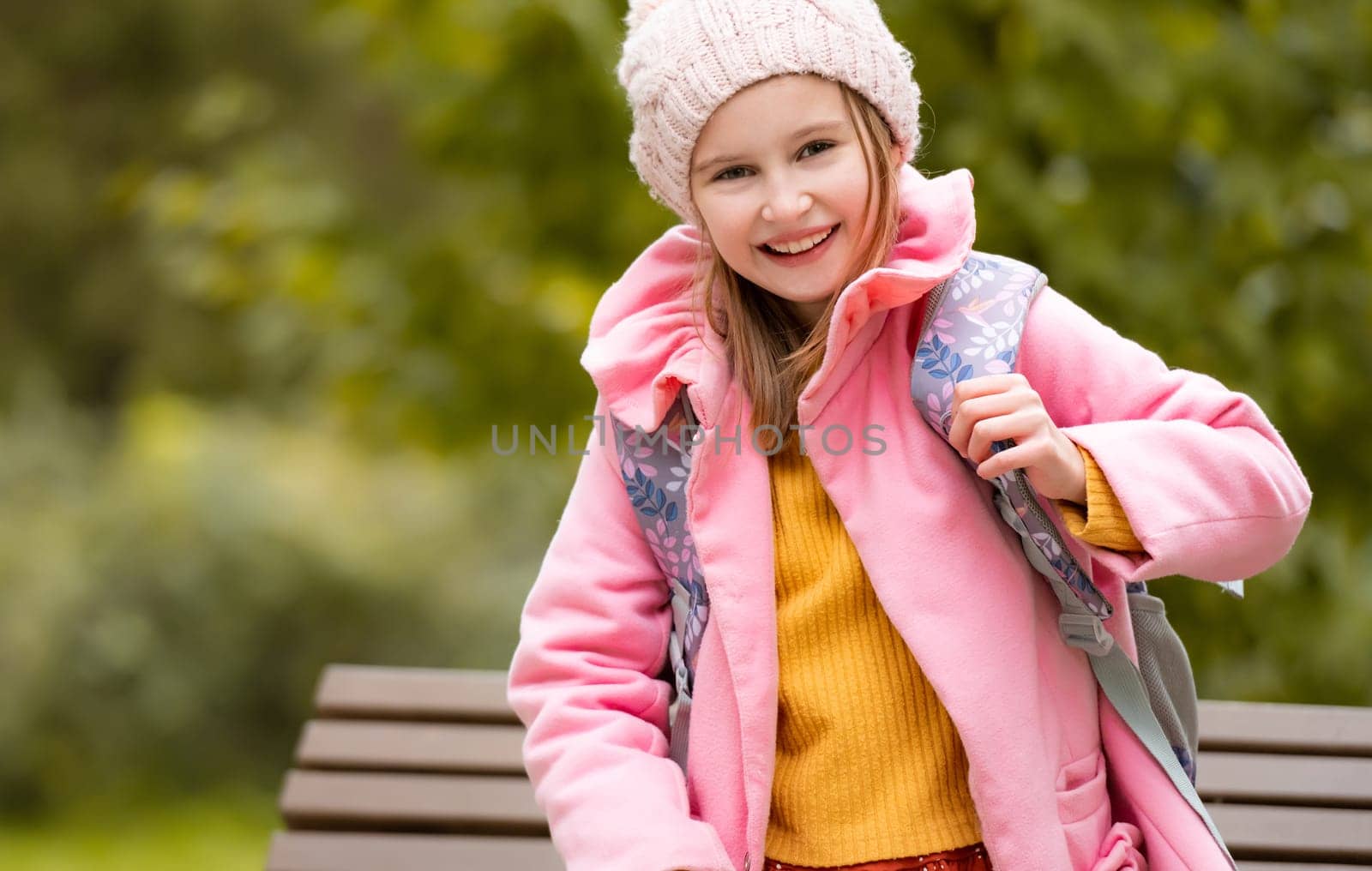 Pretty school girl kid with backpack looking at camera and smiling at autumn park. Beautiful female child portrait with blurred background