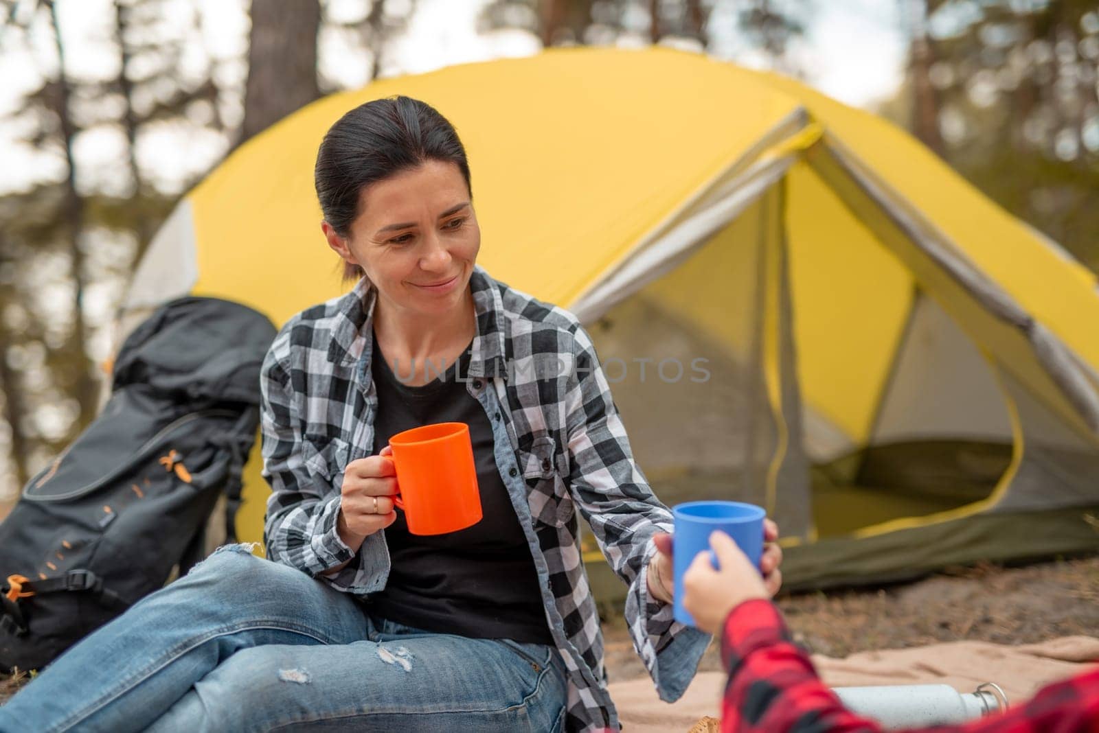 Beautiful adult girls drinking tea in camping in forest with tent on background and smiling. Young woman traveller during hiking in mountains resting