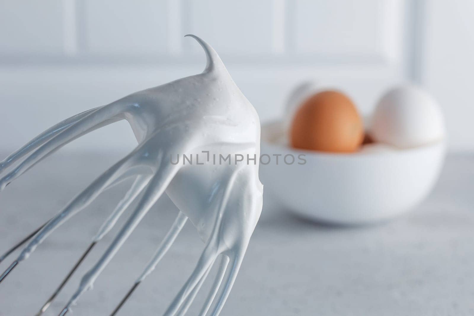 Whisked egg whites - whipped Italian meringue on a wire whisk and eggs on a gray background. copy space. by lara29