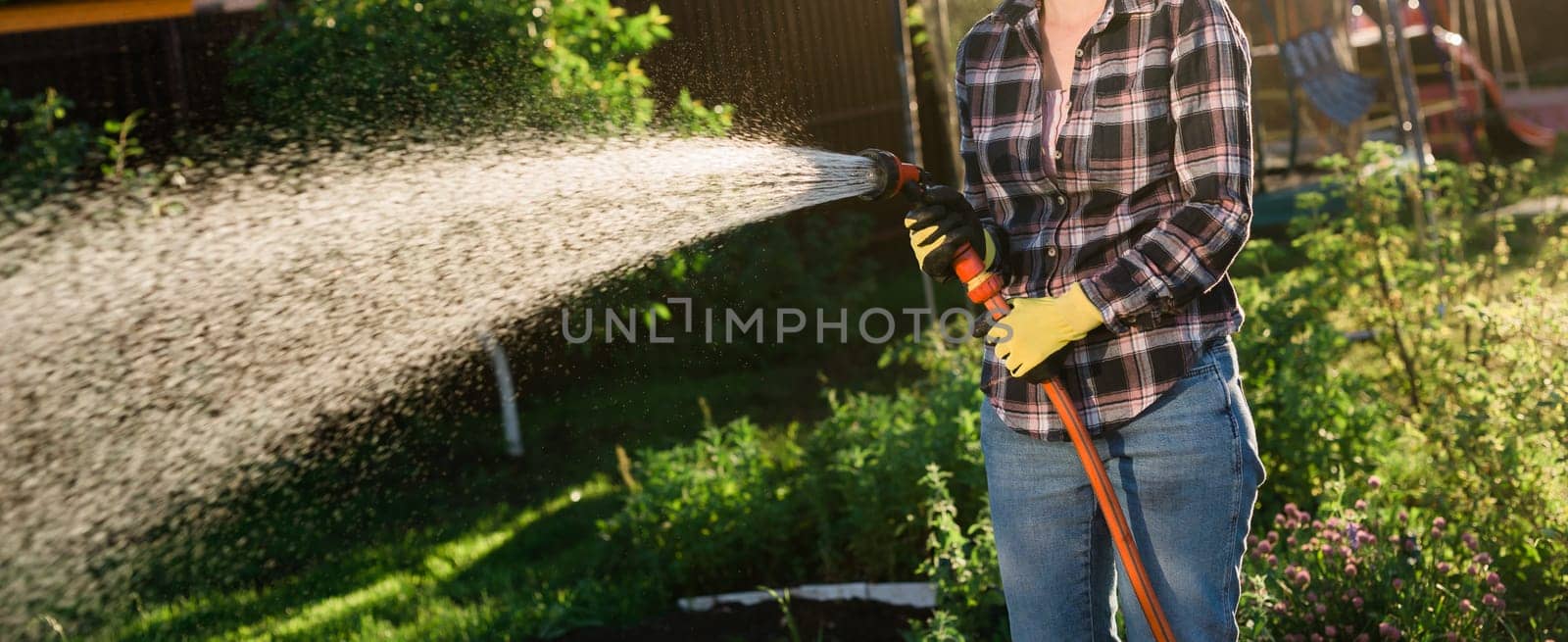 Caucasian woman gardener in work clothes watering the beds in her vegetable garden on sunny warm summer day. Concept of working in the garden