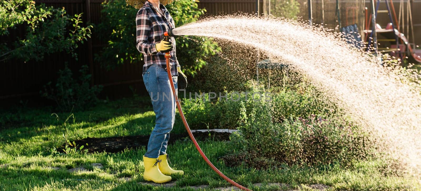 Caucasian woman gardener in work clothes watering the beds in her vegetable garden on sunny warm summer day. Concept of working in the garden