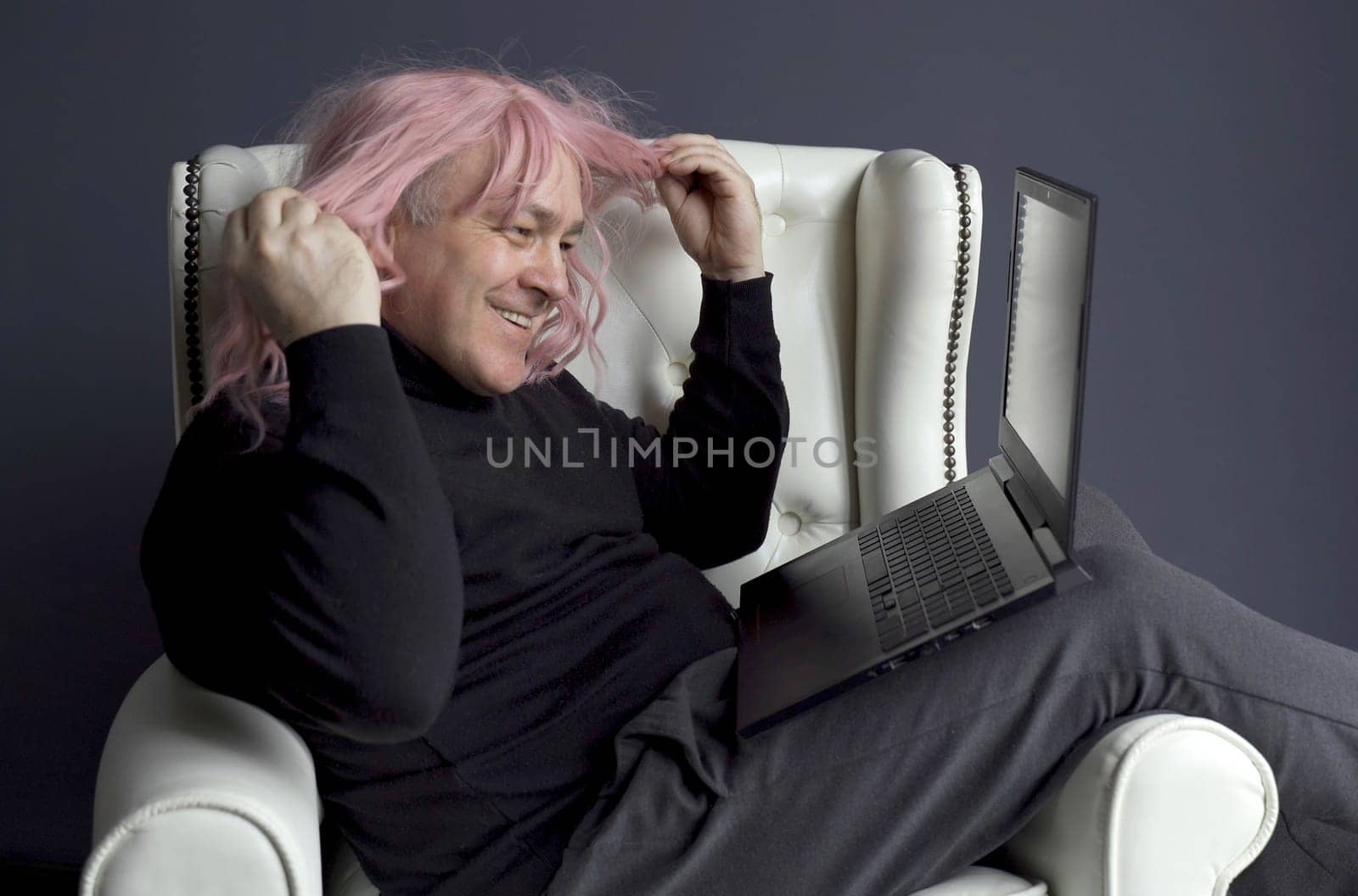 A man in a pink wig sits in a white chair and communicates via video link on a laptop. Side view