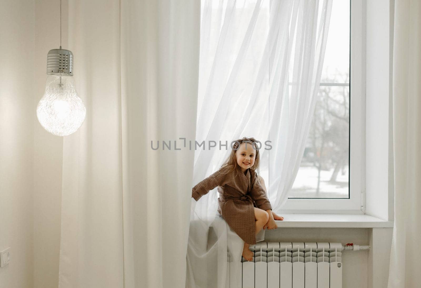 Portrait of a little girl in a brown dressing gown, who is sitting on the windowsill.