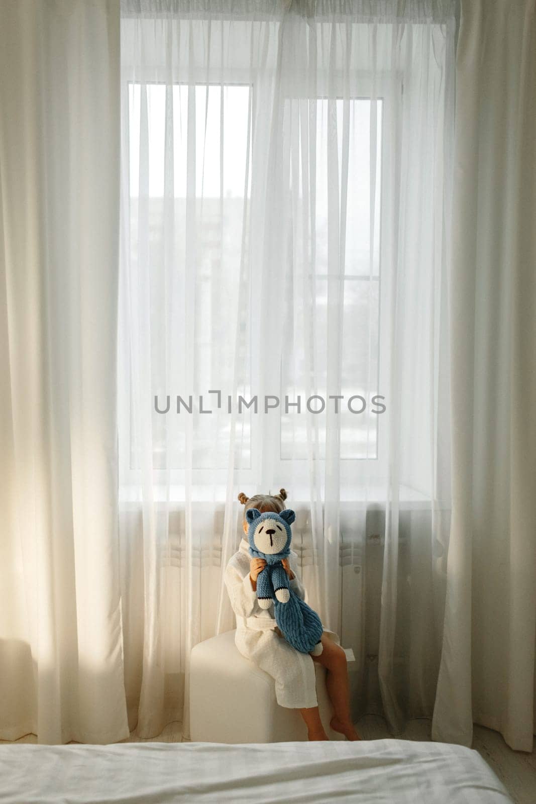 A girl in a dressing gown stands by the window and plays with a knitted toy.