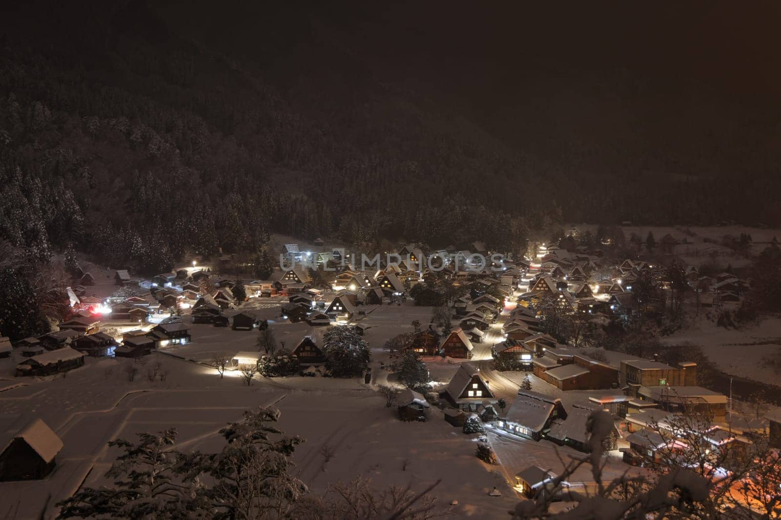 Lights from traditional mountain village pierce snow and mist at night by Osaze