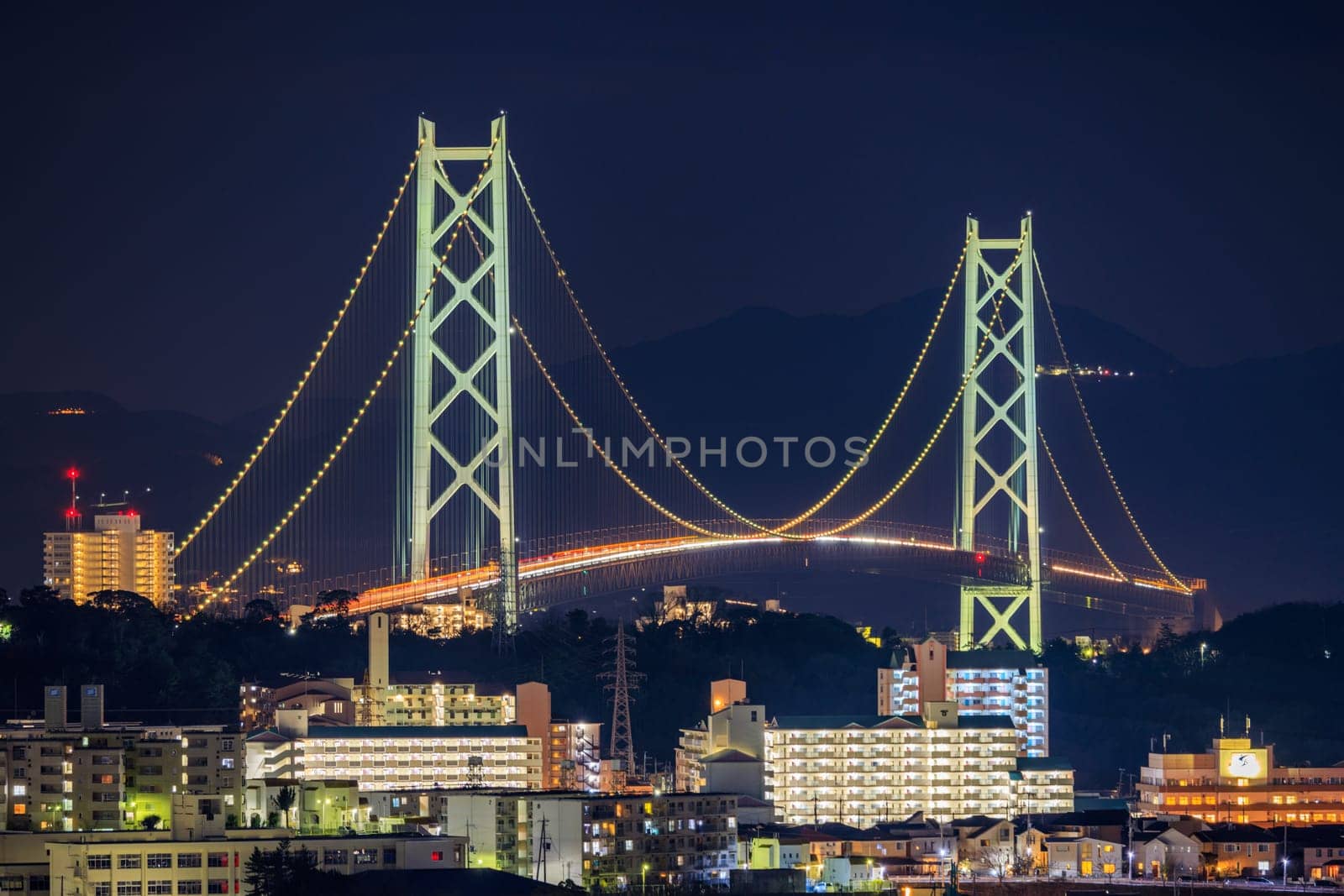 Illuminated Suspension Bridge Towers Over Apartment Buildings at Night by Osaze