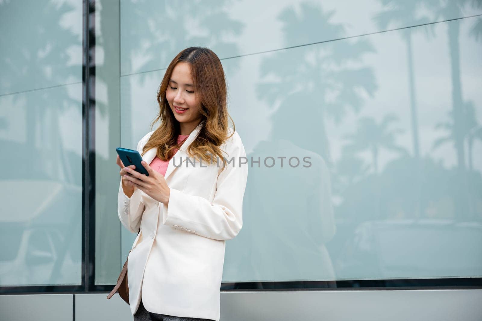 Asian businesswoman typing text on mobile phone standing outdoors street front building near office by Sorapop