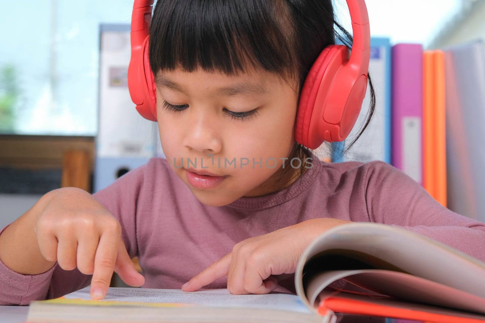 Cheerful little girl in headphones reading a book sits at the table in her room at home. Technology and hobbies for children