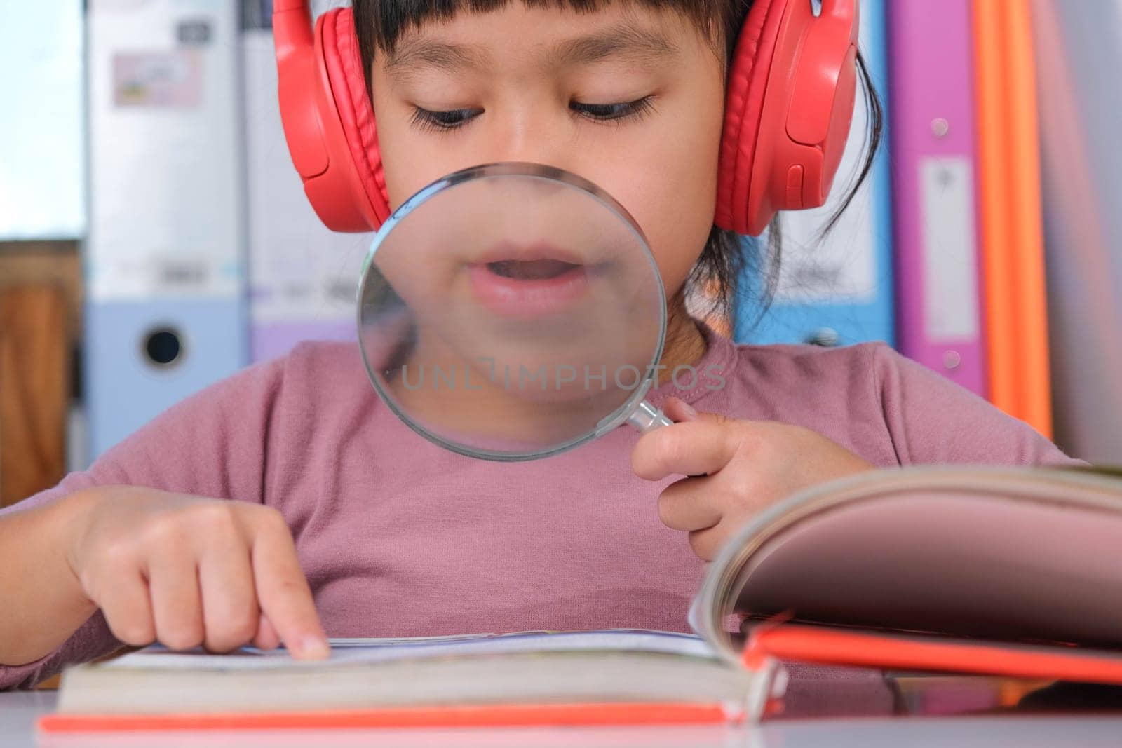 Cheerful little girl in headphones reading a book with a magnifying glass sitting at the table in her room at home.