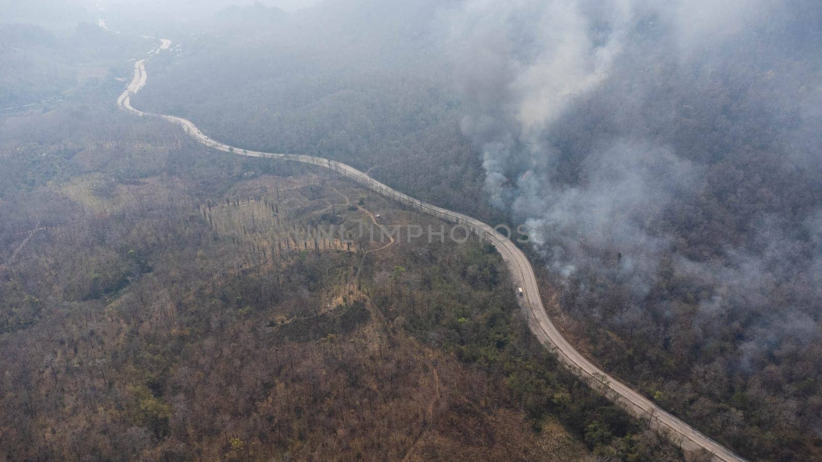 Aerial drone view of a wildfire burning through a forest area, fills the sky with dark smoke in the woods near the edge of the highway. Burning Forest. Air pollution concept by TEERASAK