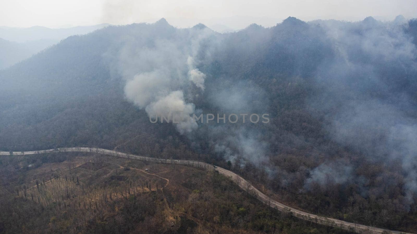 Aerial drone view of a wildfire burning through a forest area, fills the sky with dark smoke in the woods near the edge of the highway. Burning Forest. Air pollution concept by TEERASAK