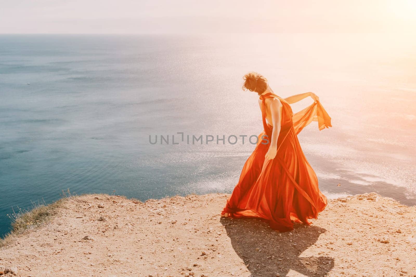 Side view a Young beautiful sensual woman in a red long dress posing on a rock high above the sea during sunrise. Girl on the nature on blue sky background. Fashion photo.