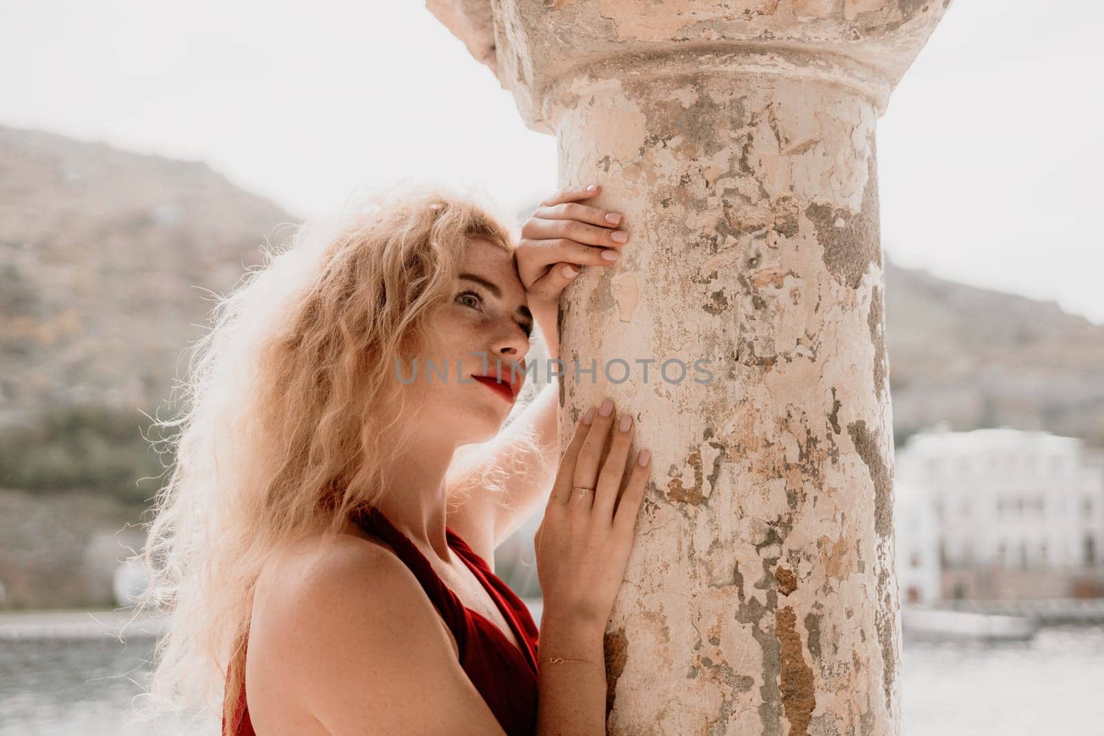 Redhead woman portrait. Curly redhead young caucasian woman with freckles looking at camera and smiling. Cute woman close up portrait in a red long dress posing on sea and travel city background by panophotograph