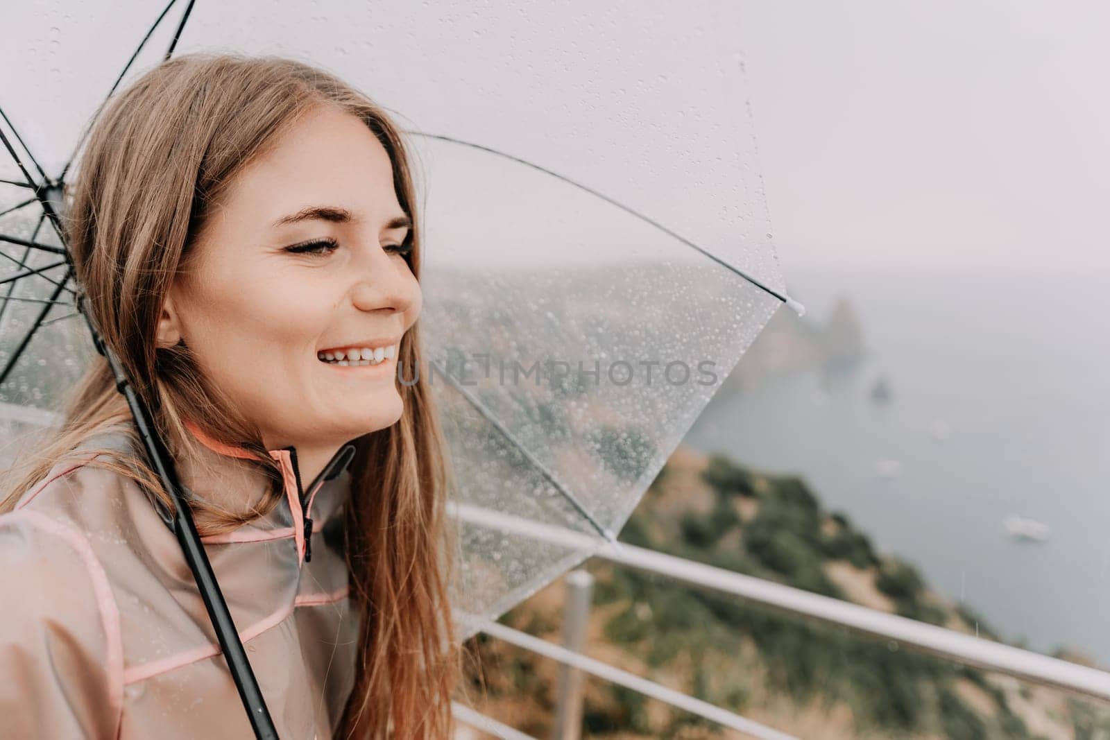 Woman rain park. Happy woman portrait wearing a raincoat with transparent umbrella outdoors on rainy day in park near sea. Girl on the nature on rainy overcast day