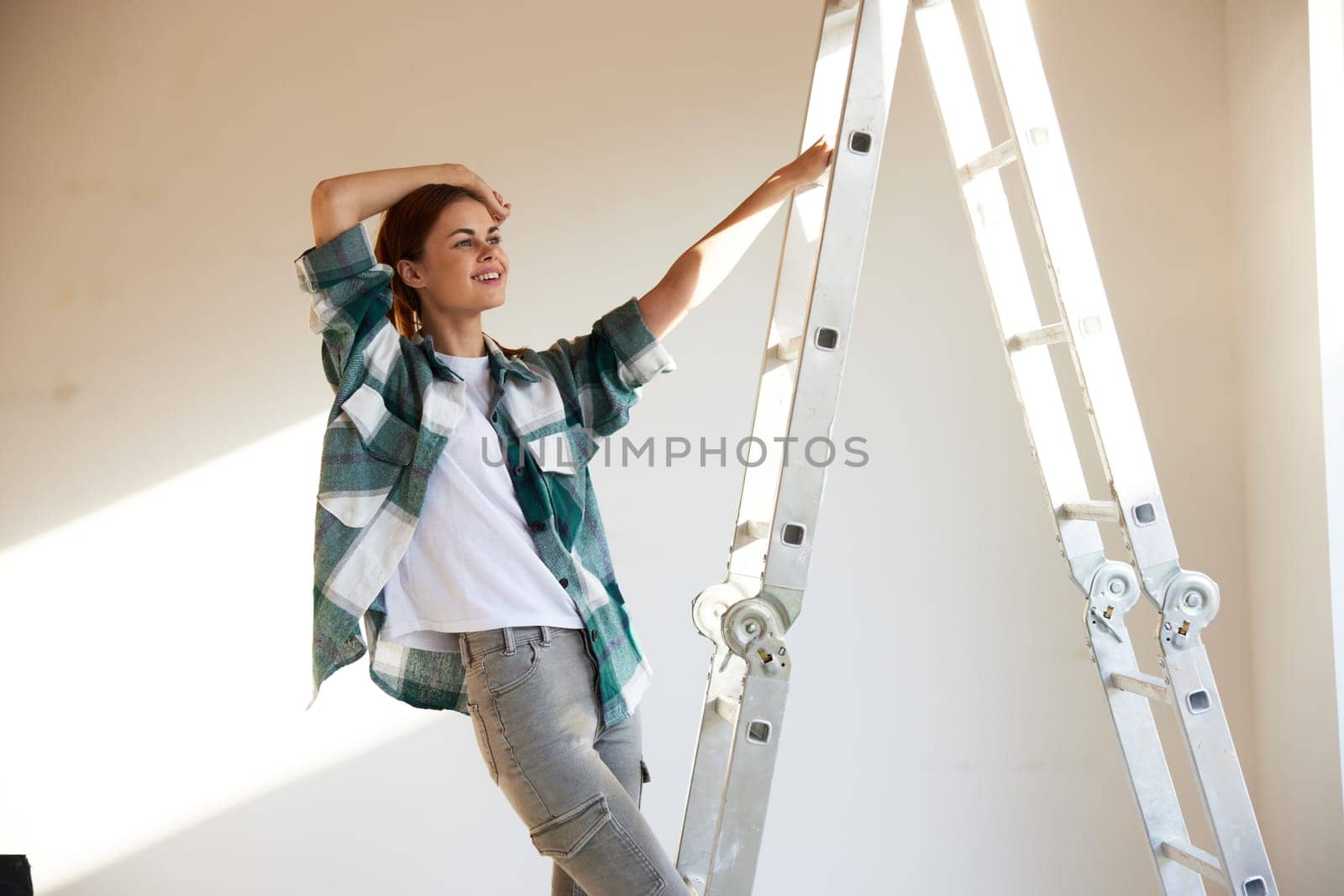 a laughing woman stands on a construction ladder and joyfully raises her hand as a sign of the end of the repair. High quality photo