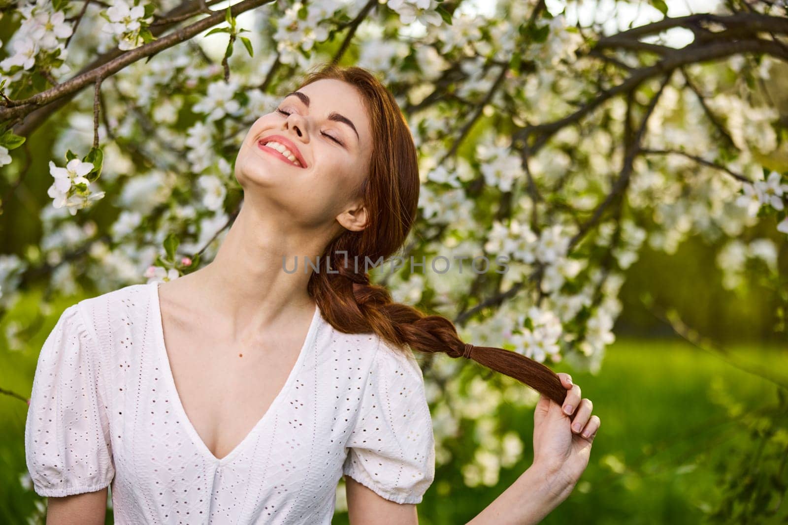 portrait of a joyful woman in a light dress against the background of a flowering tree, holding herself by her braid by Vichizh