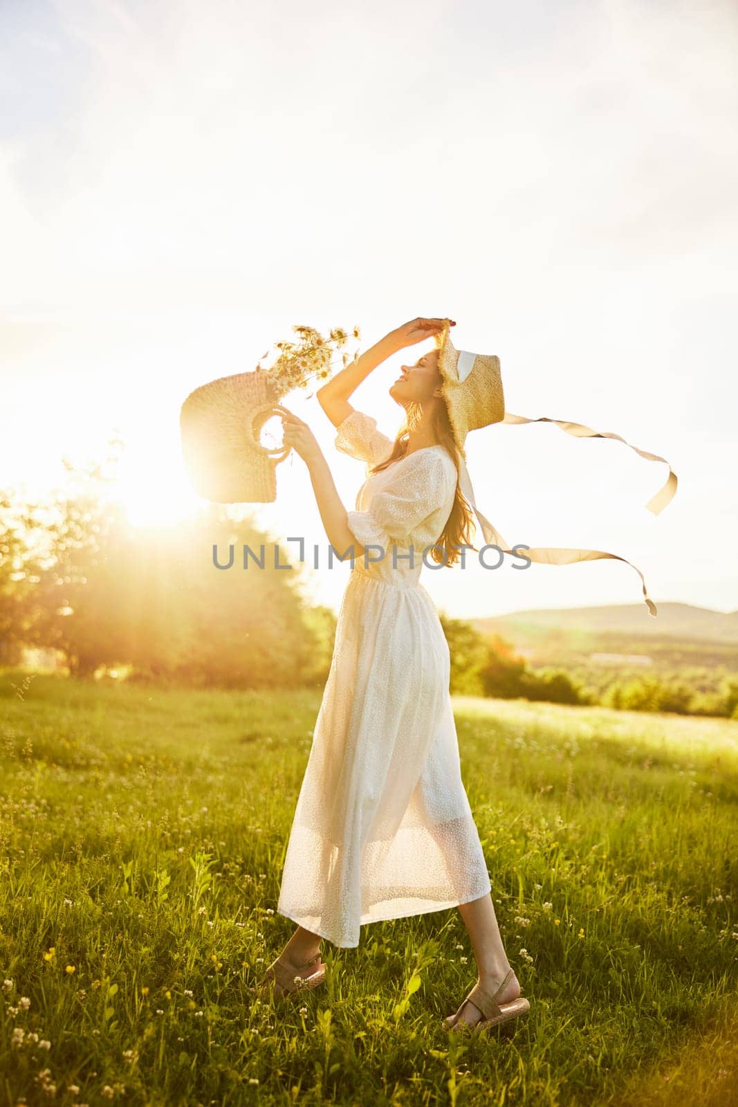 a woman in a white dress with a basket of daisies and a straw hat stands in a field during sunset in a light breeze. High quality photo