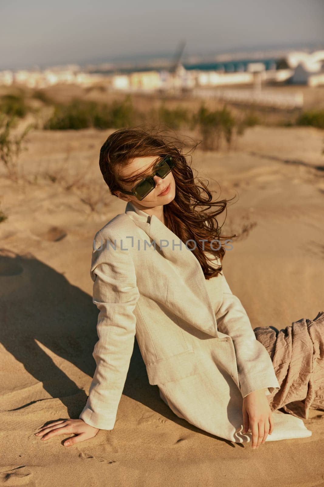 a woman in a stylish jacket and dark glasses sits on the sand relaxing by Vichizh
