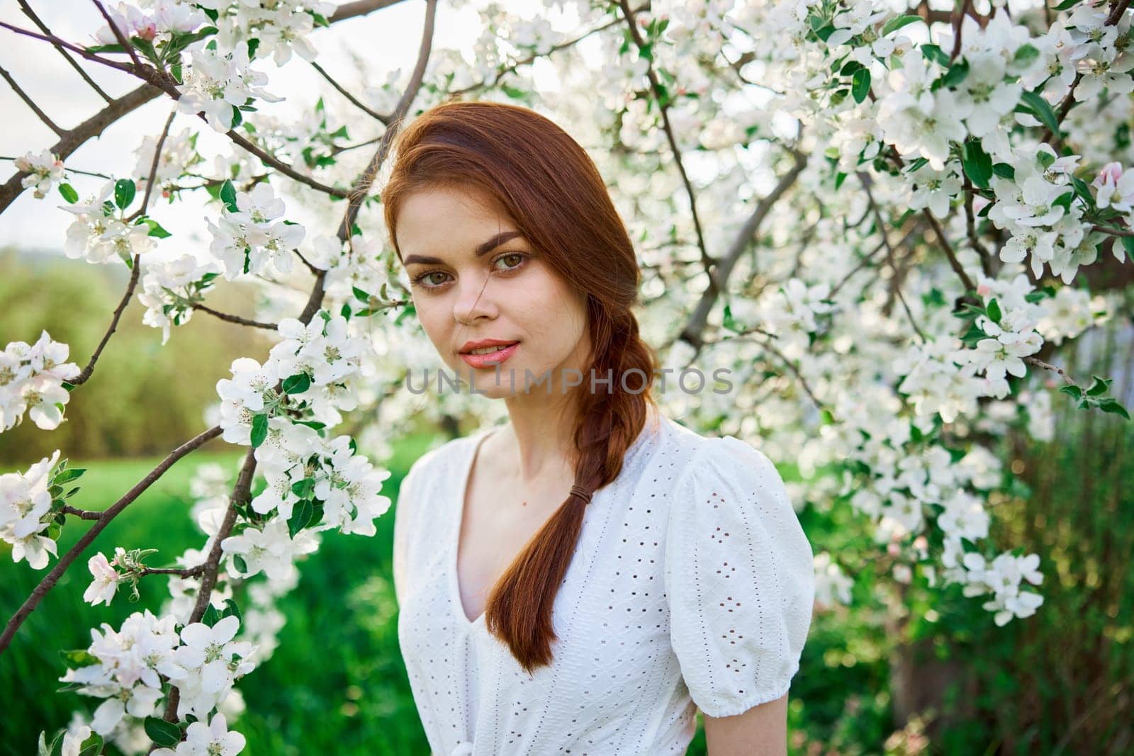 beautiful red-haired woman in a white dress smiling stands near a flowering tree by Vichizh