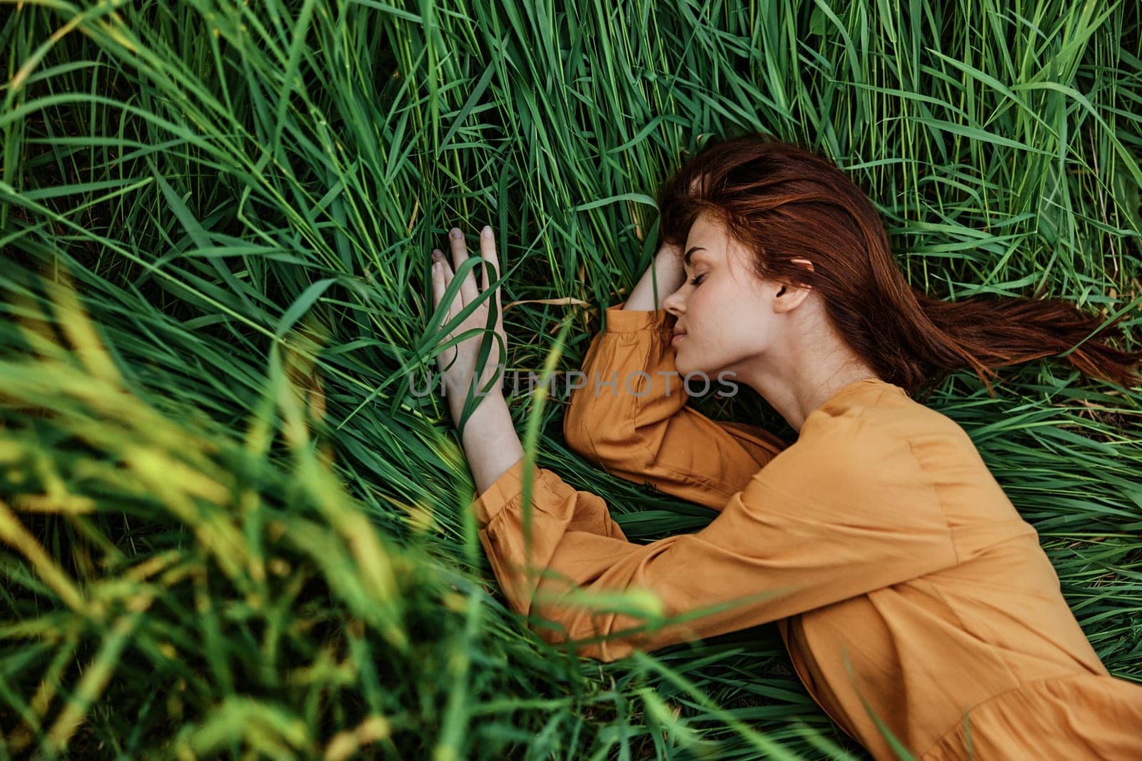 a close horizontal photo of a pleasant woman in a long orange dress resting lying in the tall grass with her eyes closed in sunny weather at sunset with her arms outstretched. Street photography,. High quality photo