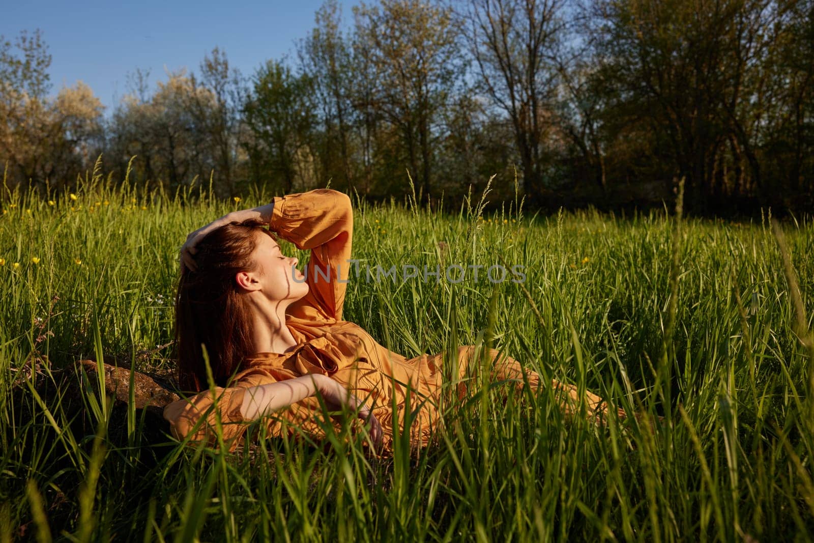 a beautiful, stylish woman lies in a long orange dress in the tall green grass illuminated by the sun rays with her eyes closed, holding her hand near her head and looking away by Vichizh