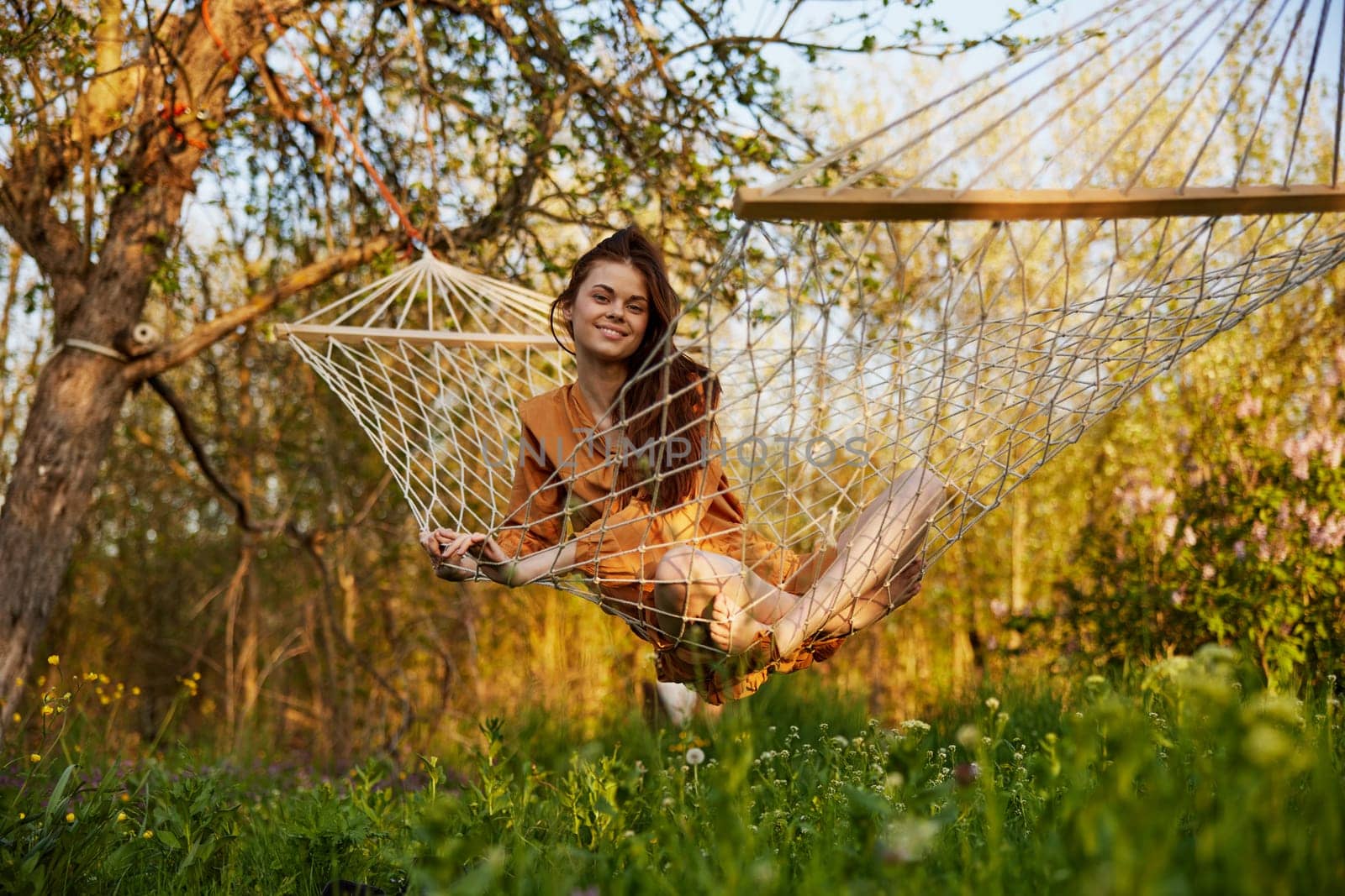a happy woman in a long orange dress is resting sitting in a hammock at the dacha, smiling pleasantly at the camera, illuminated by the summer sun during sunset by Vichizh
