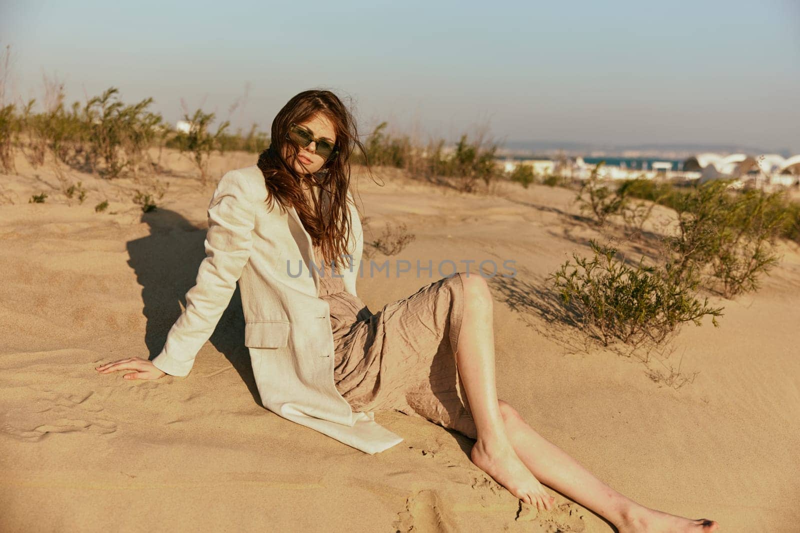 a woman in light clothes and black sunglasses sits on the sand in windy weather by Vichizh