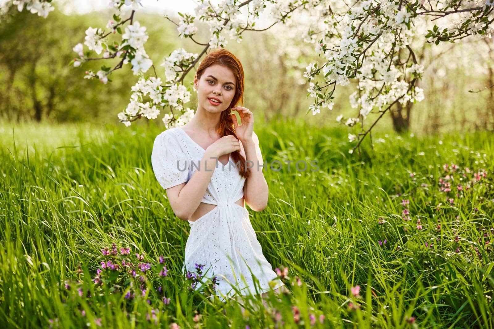 a young woman in a light summer dress stands near a tree with flowers and smiles looking at the camera. High quality photo