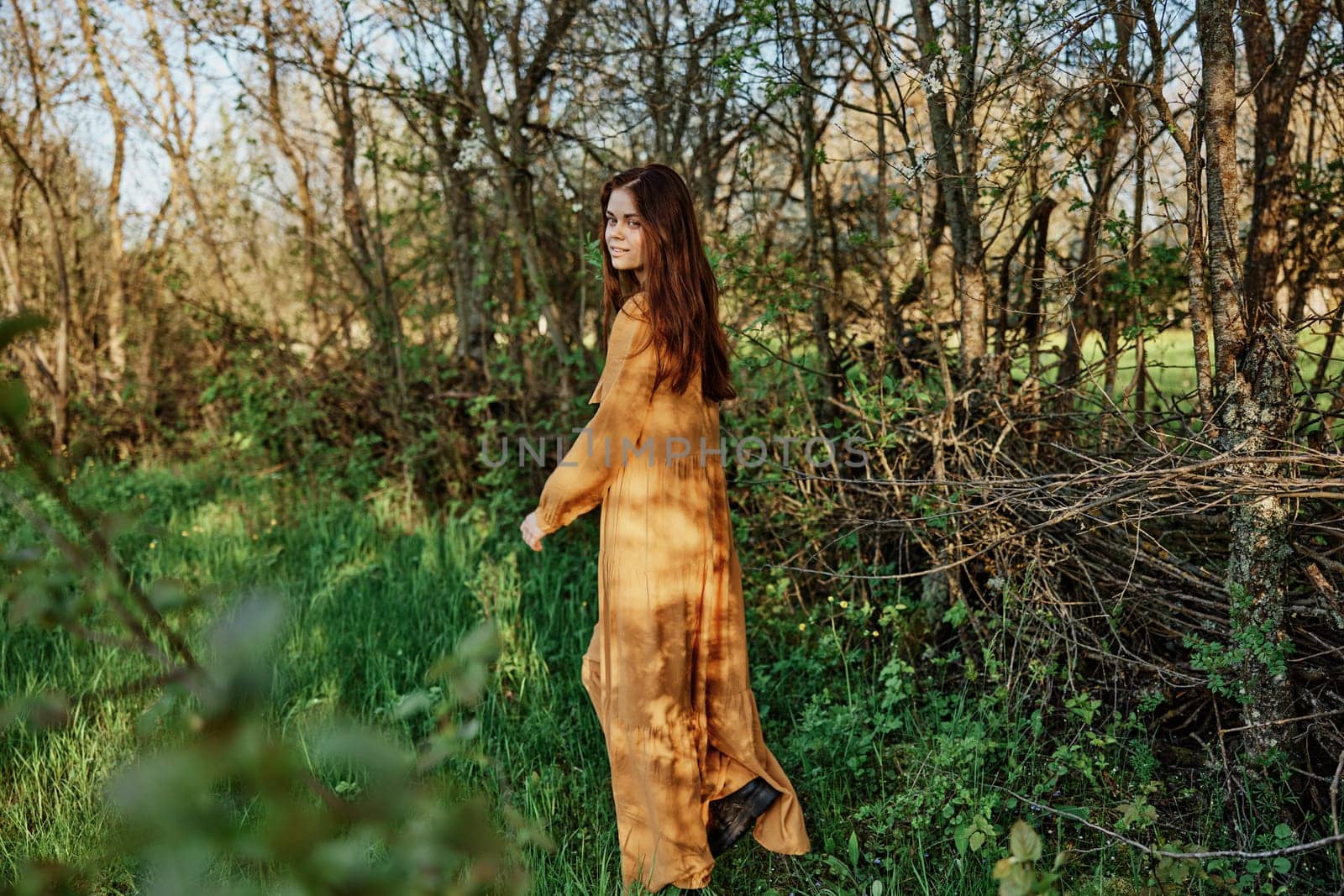 a beautiful woman with long hair is standing in the countryside in a long orange dress smiling pleasantly and looking away. High quality photo