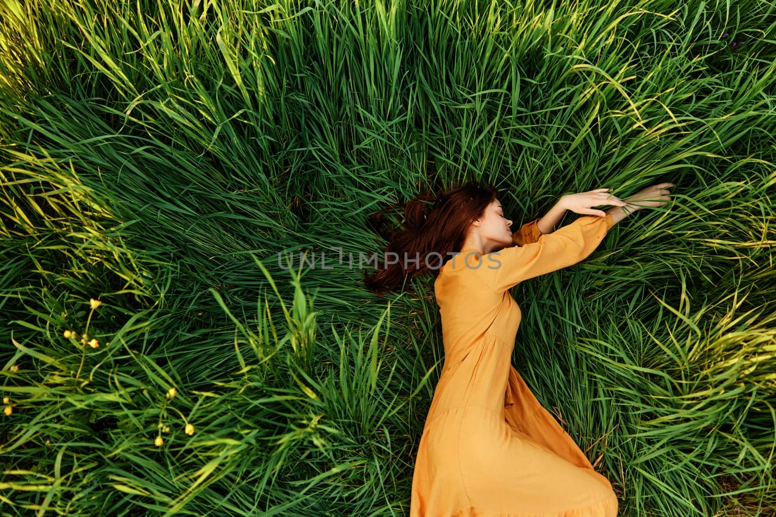 an elegant woman lies in the tall green grass in a long summer orange dress and relaxed posing with her hands enjoying nature and sunny weather with her eyes closed. High quality photo