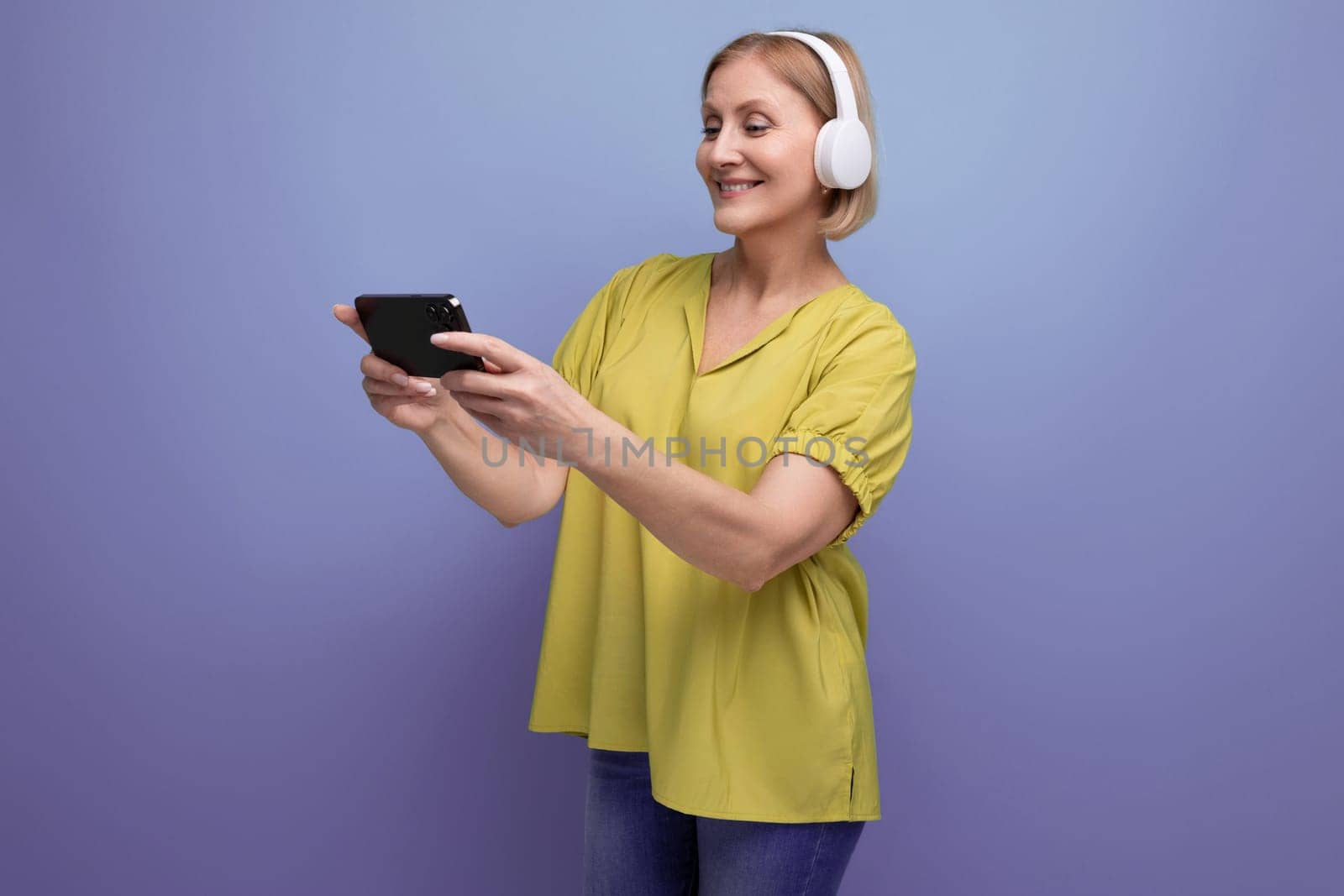 blonde 50s woman watching broadcast on smartphone using headphones.