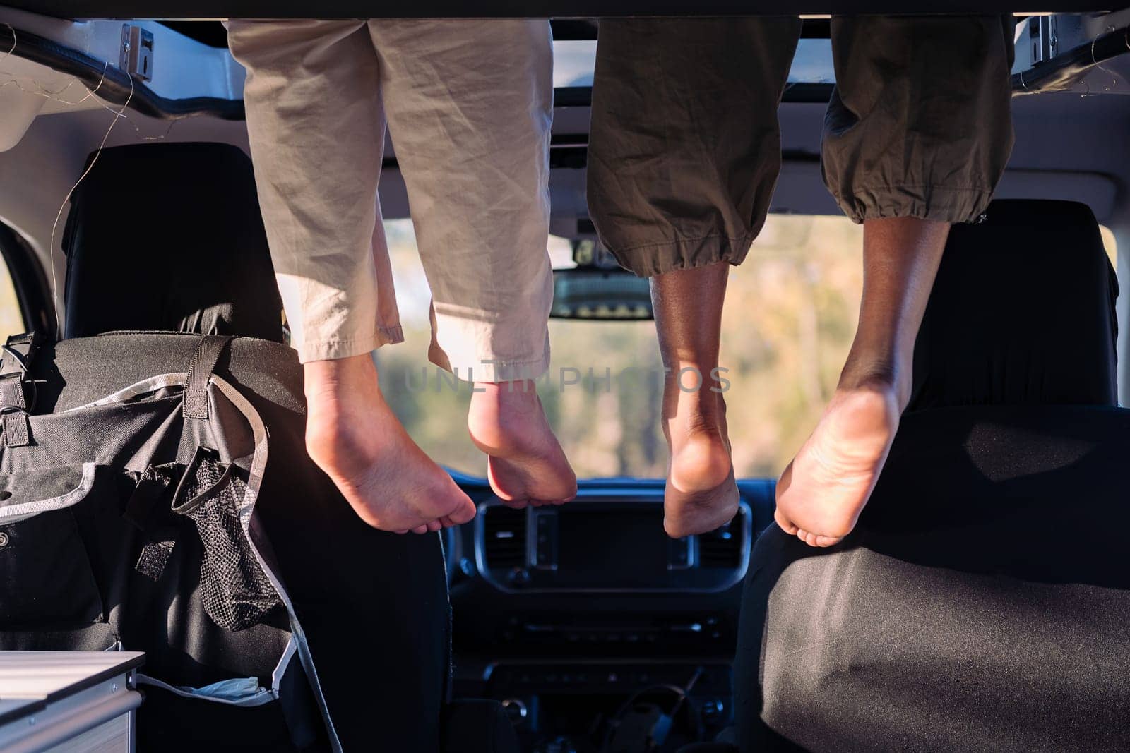 two unrecognizable women relaxing with feet dangling off the elevated bed of a camper van, concept of road trip adventure with best friend and van life relaxation