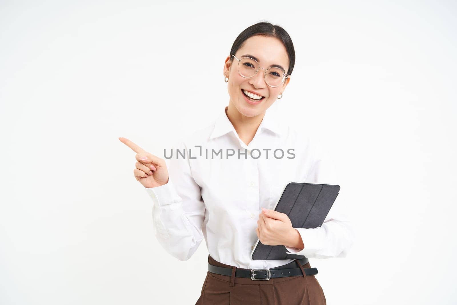 Young professional, businesswoman in glasses holding digital tablet, pointing finger left, showing business advertisement, standing over white background by Benzoix