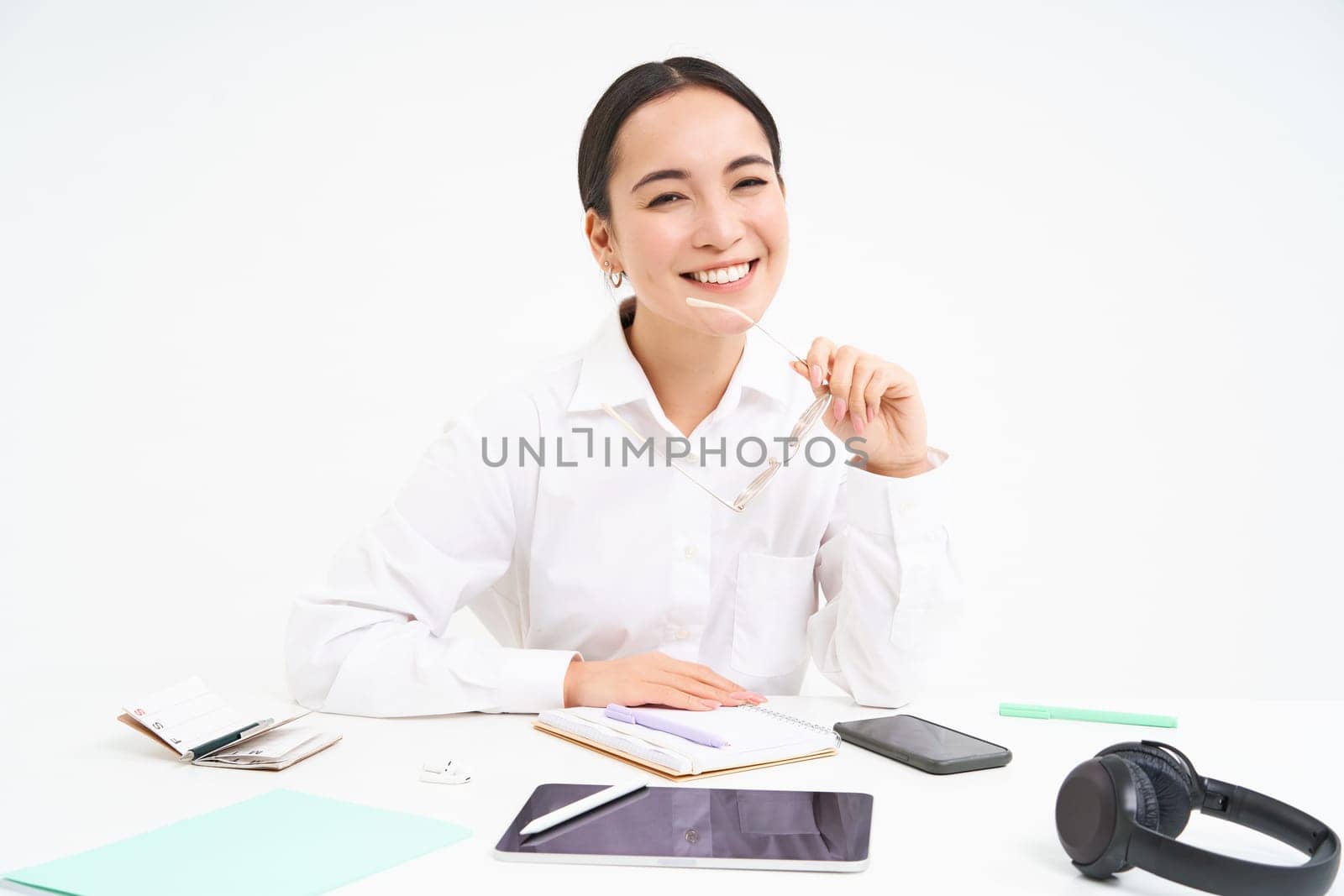 Portrait of successful businesswoman, office manager at workplace, sits with digital tablet and her working documents, wears glasses, smiles at camera.