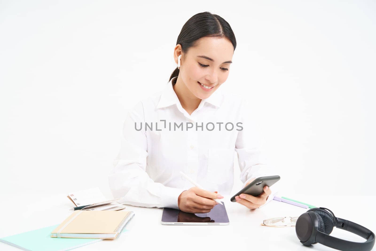 Image of businesswoman sits in office with digital tablet and headphones, looks at smartphone, checks clients messages, calling employee, white background.