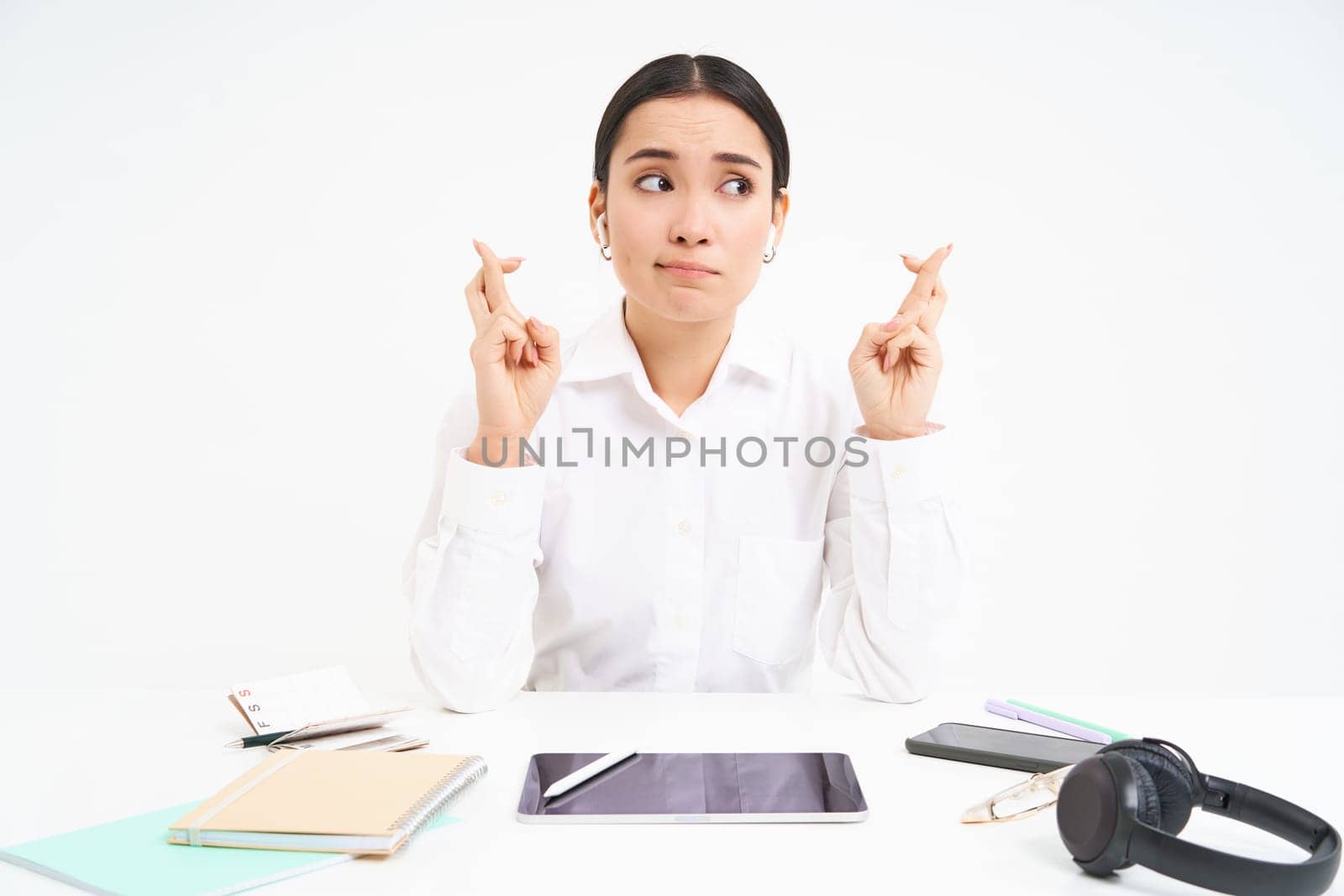 Hopeful asian businesswoman, cross fingers for good luck, sits in workplace office and makes wish, white background.