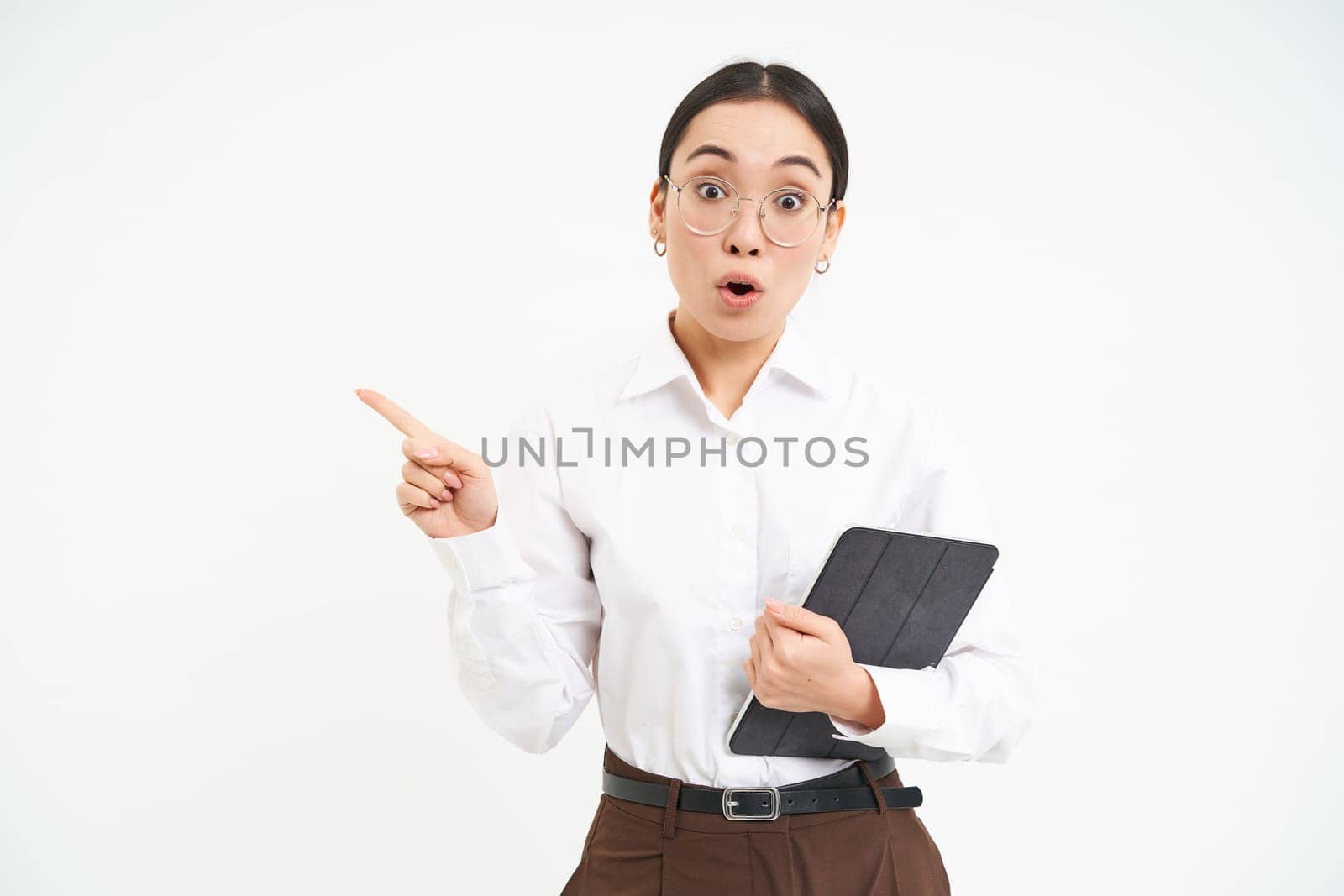 Portrait of korean businesswoman looks surprised, points left and looks amazed, holds digital tablet, stands over white background.
