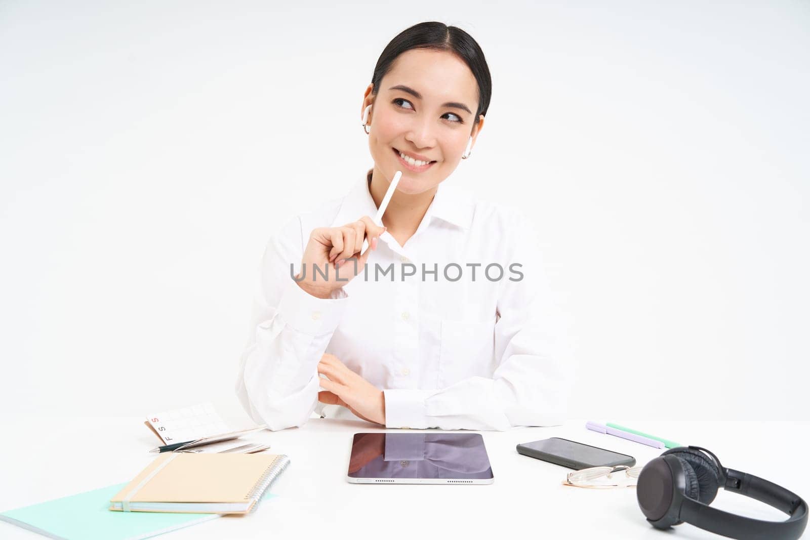 Young asian saleswoman in her office, working on digital tablet, has notebooks and headphones on her table, white background.