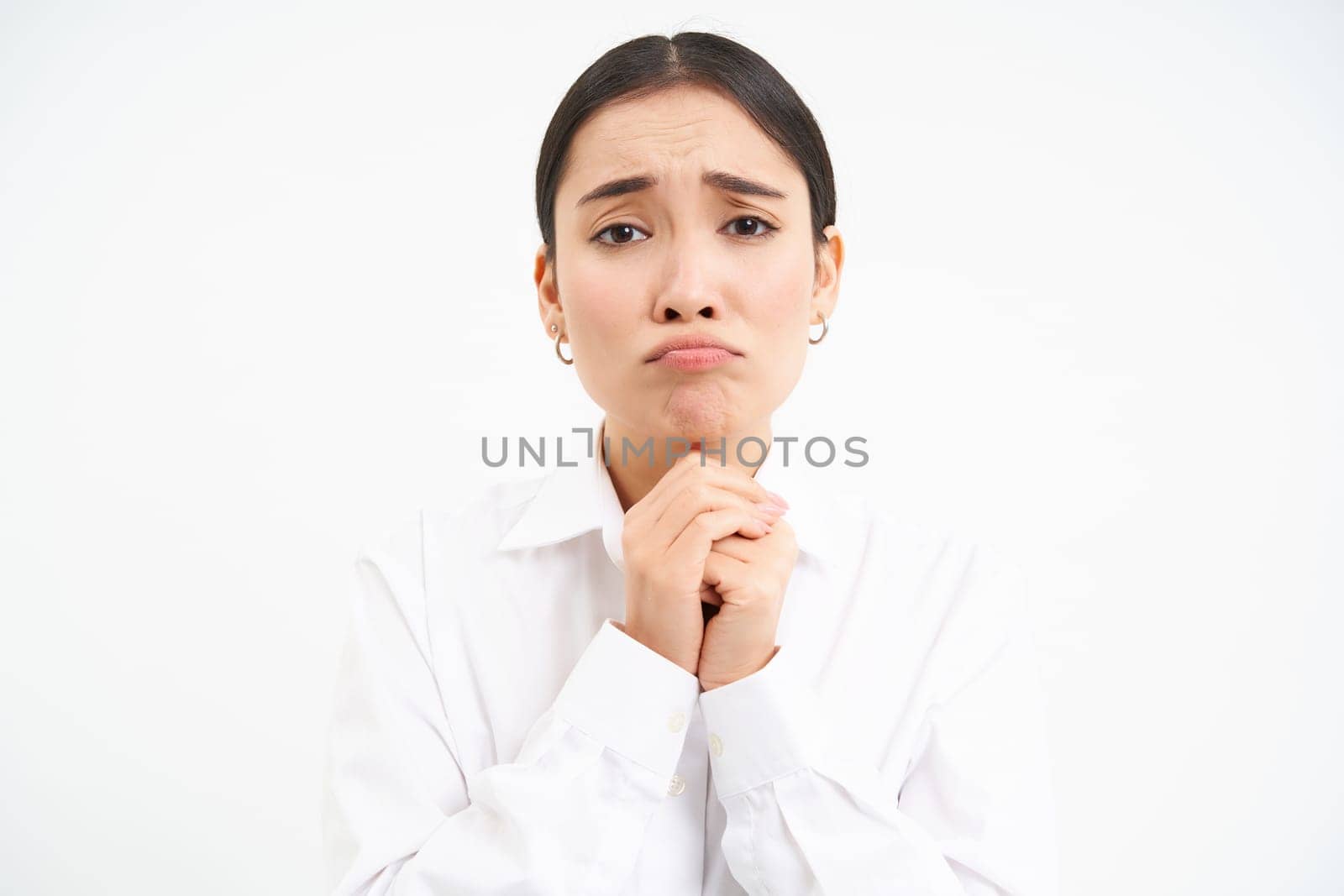 Upset young saleswoman, asian woman holds hands in pray, begs you, asks for favour with sad face, pleading, standing over white background by Benzoix