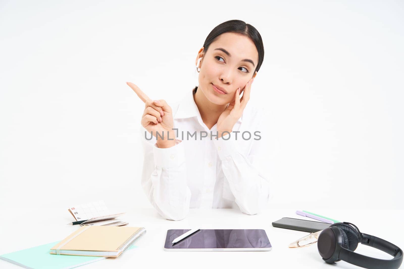 Business and workplace. Young asian woman in office, sits with digital tablet, points finger left, shows banner, white background.