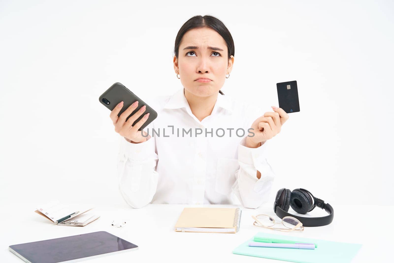 Portrait of asian woman sits at table, holds credit card and smartphone, looks upset, disappointed by prices, white background.