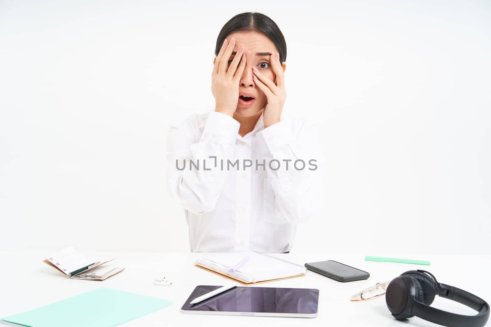 Image of asian office lady shuts her eyes embarrassed, sees something scary, sits at workplace, white background.