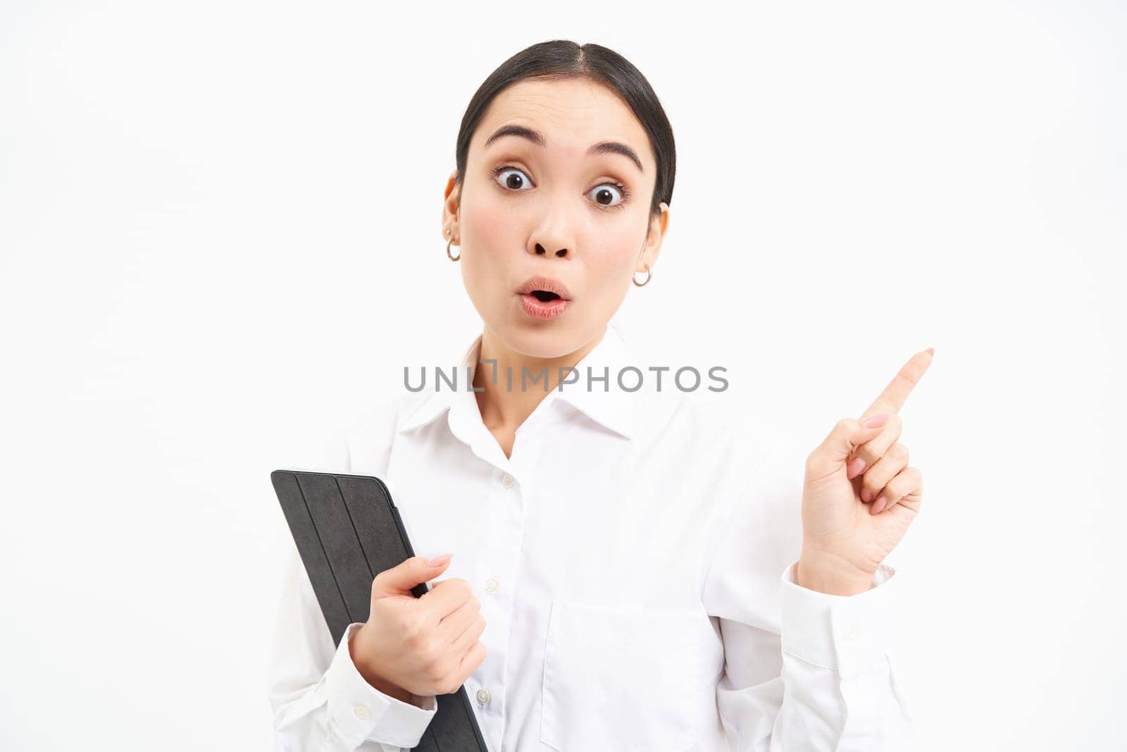 Image of young professional woman, pointing finger right, holding digital tablet, showing advertisement campaign, isolated on white background.