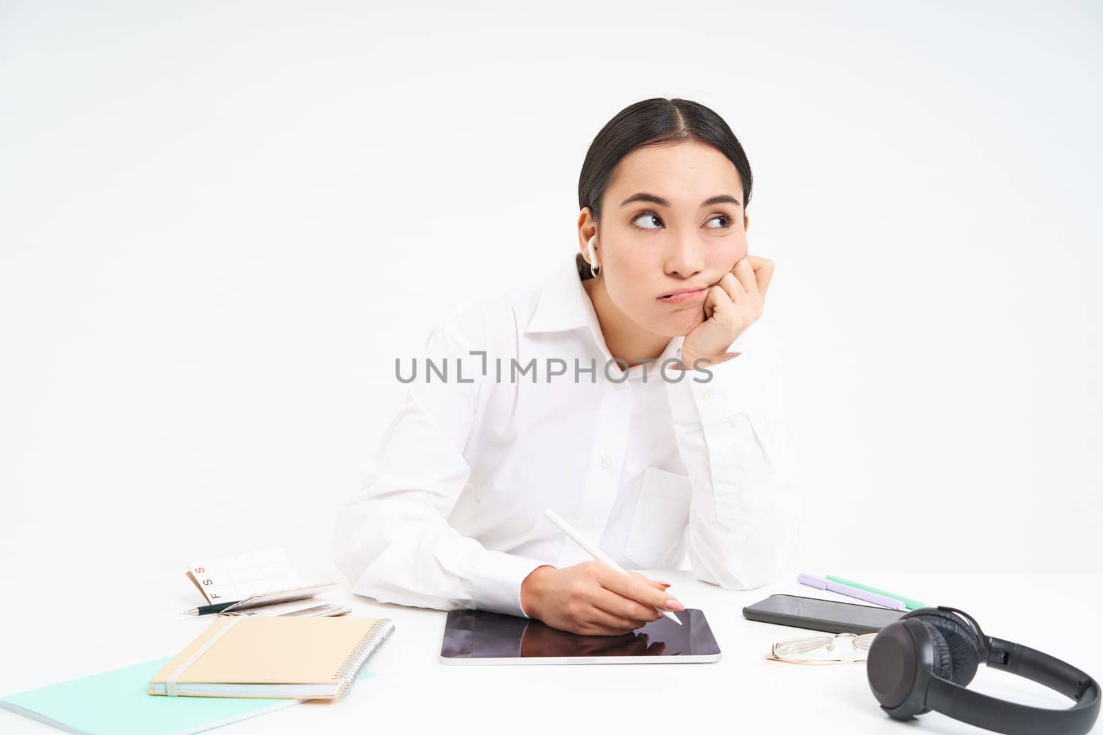 Tired and bored korean woman in office, employee sits with digital tablet, listens music in earphones and looks tired, white background.