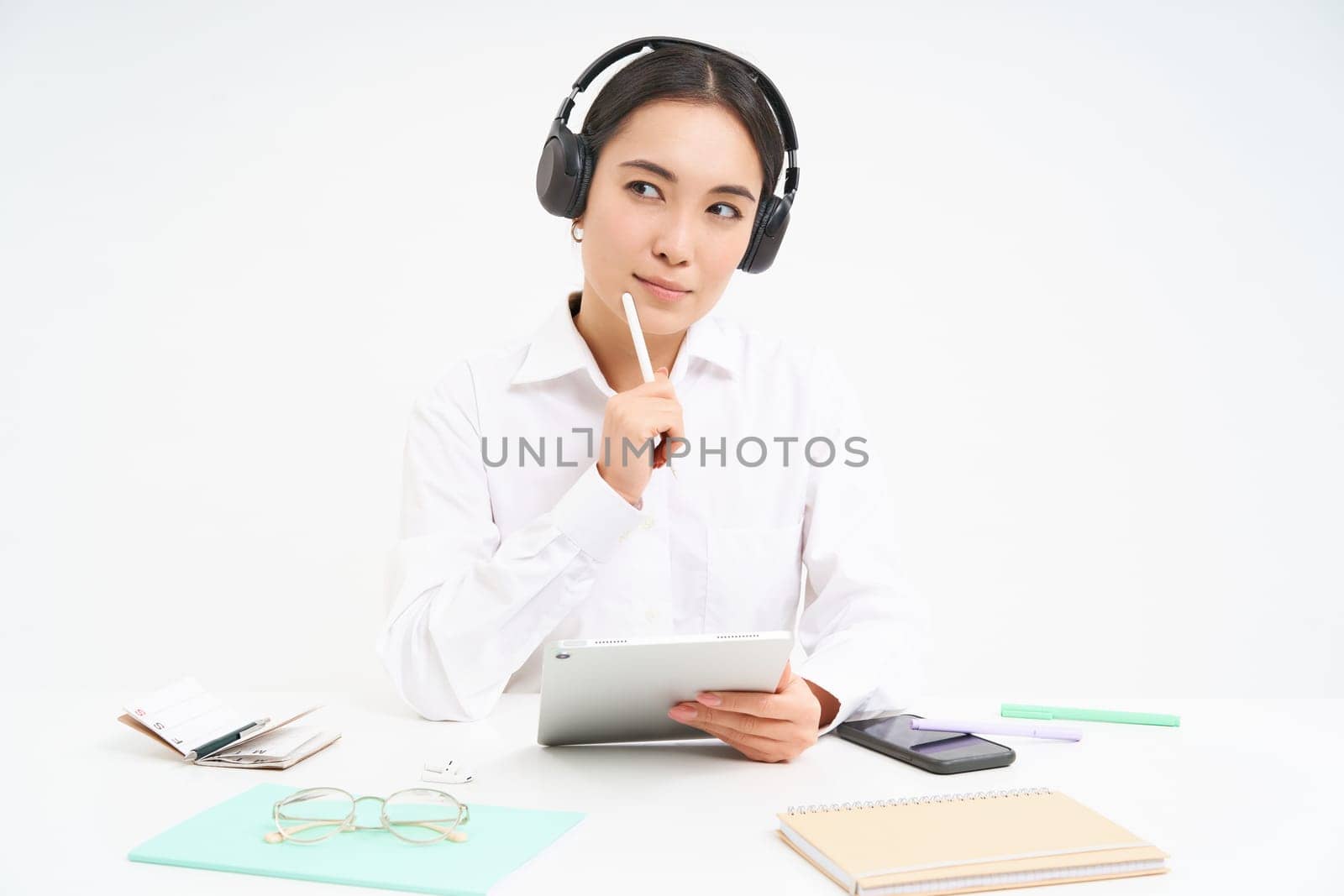 Portrait of professional woman working, listening podcast or course in headphones, holding digital tablet, sitting over white background.