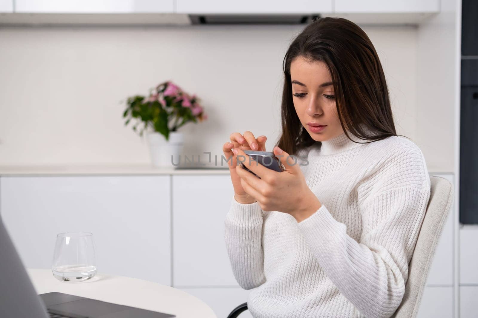 Young woman looking at the screen of a smartphone sitting in the kitchen. Female texting with her boyfriend concept.